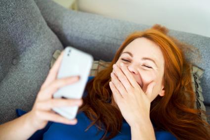 Femme allongée sur le canapé à l'aide de téléphone intelligent, souriant derrière la main